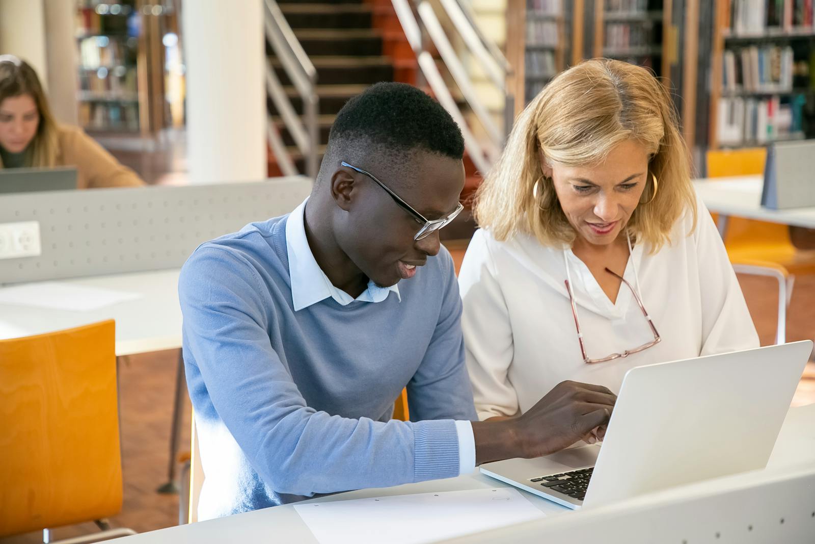 Happy young adult male student wearing eyeglasses with middle-aged female tutor in a library